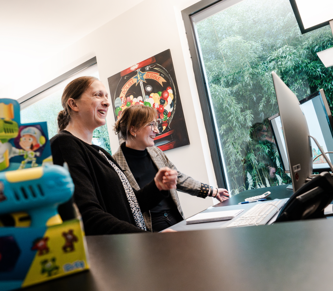 Zwei Frauen lächeln und schauen auf einen Computerbildschirm, während eine farbige Spielzeugfigur im Vordergrund zu sehen ist. Im Hintergrund hängt ein Bild mit Bonbons.