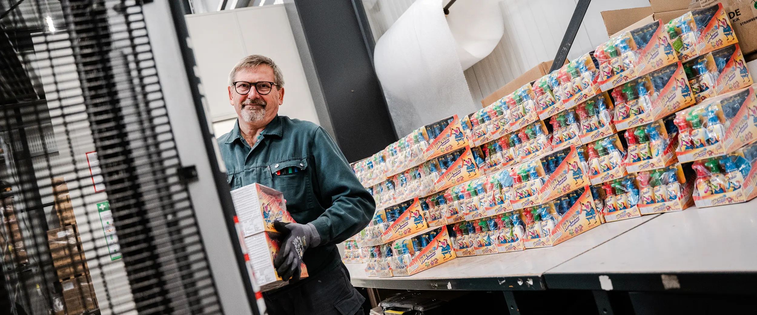 Mann mit Arbeitshandschuhen und einer Brille lächelt, während er Kartons mit Spielzeug verräumt