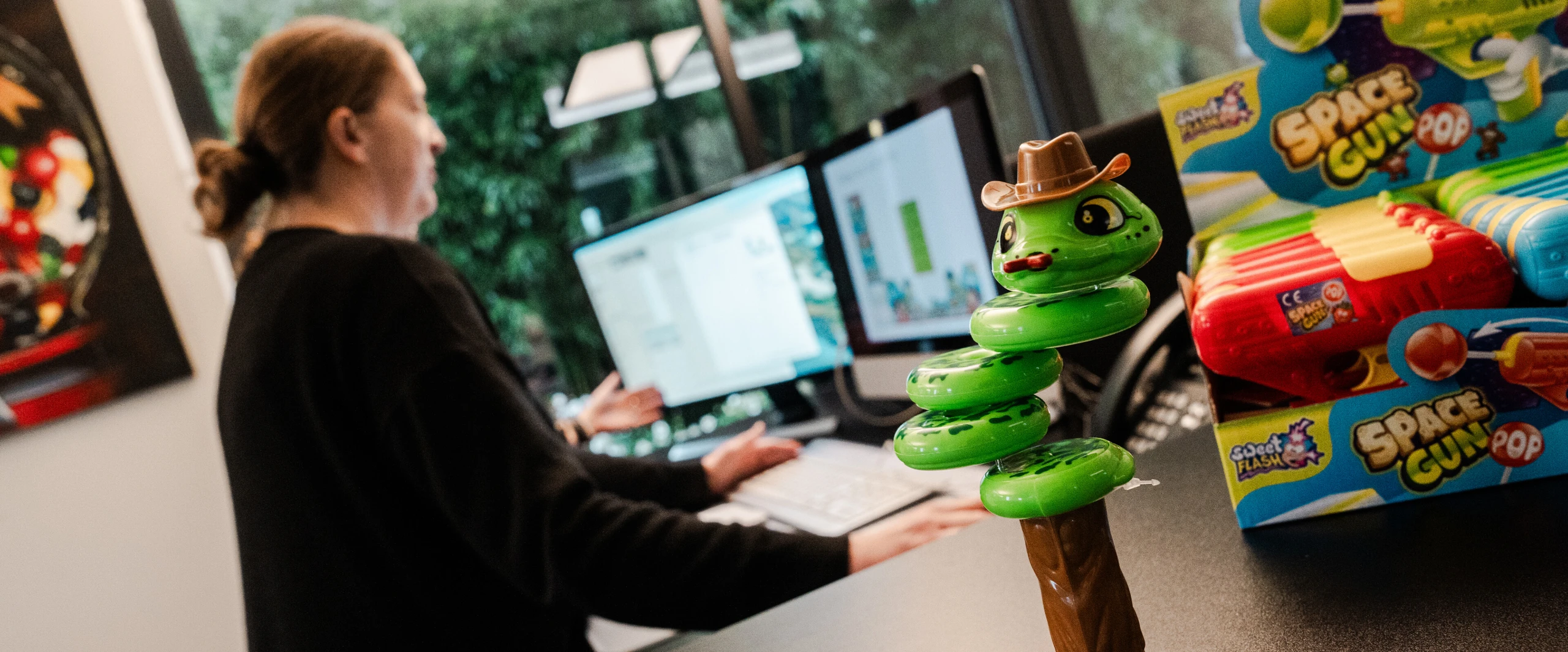 Eine Person arbeitet an einem Computer, während ein bunter Spielzeug-Schnappschlange im Vordergrund steht. Im Hintergrund sind Spielzeug-Waffen zu sehen.
